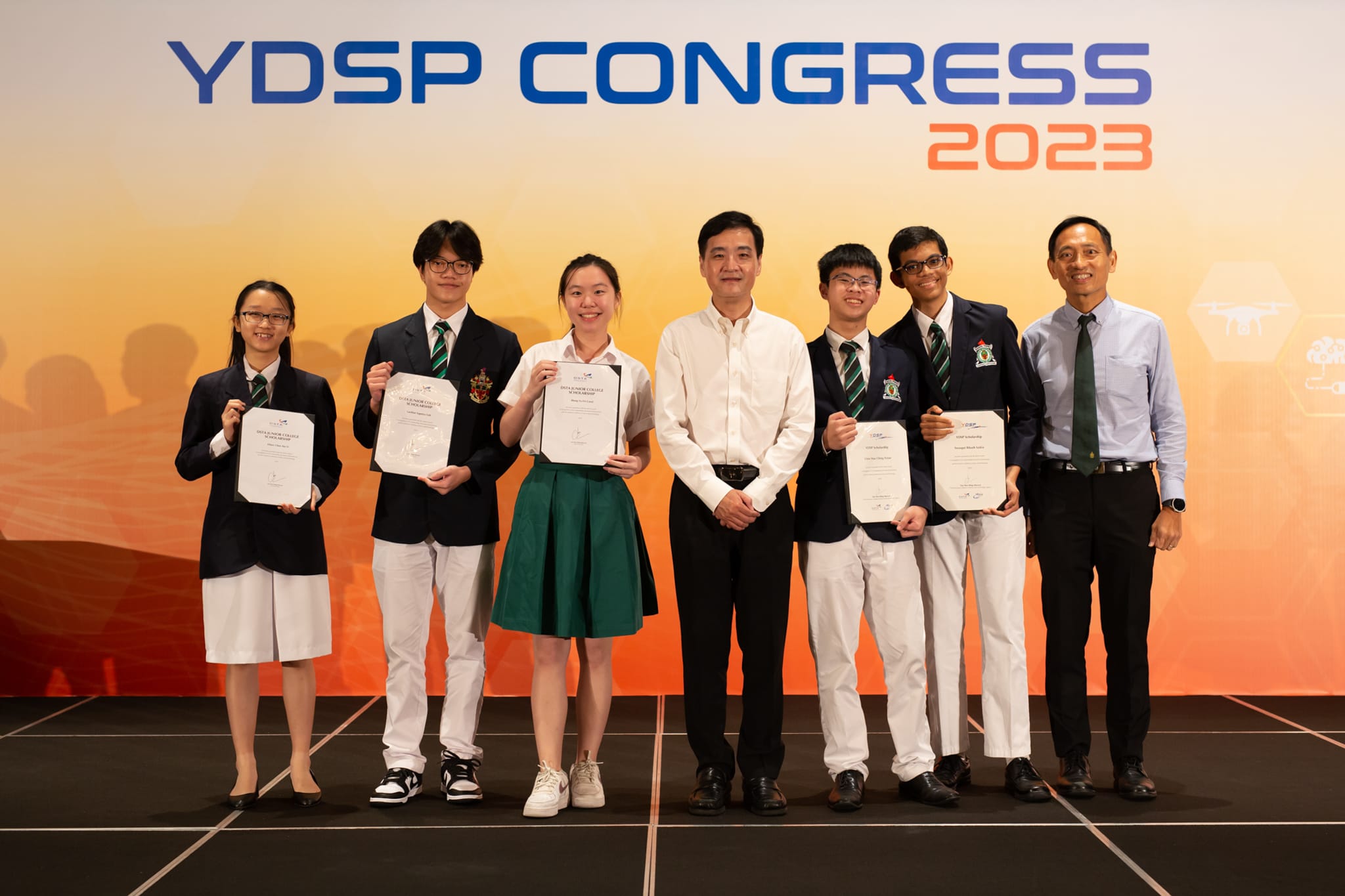 Seven people pose on stage at YDSP Congress 2023, with five students holding certificates and two adults standing beside them.